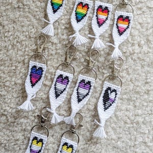May include: A collection of white keychains with silver clasps, each featuring a colorful, embroidered heart design. The hearts display various LGBTQ+ pride flags, including rainbow, lesbian, and non-binary flags. Each keychain has a white tassel.