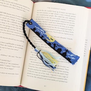 Puede incluir: Un marcapáginas tejido rectangular con un patrón de noche estrellada en azul, negro y amarillo. El marcapáginas tiene un cordón negro trenzado unido a un lazo en la parte superior y una borla multicolor de hilos azules, amarillos y negros.