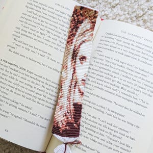 Puede incluir: Un marcapáginas de macramé rectangular con un retrato tejido en tonos blanco, beige y marrón oscuro. El diseño muestra un rostro con patrones de nudos intrincados. El borde inferior está rematado con un fleco de borla blanco y marrón oscuro.
