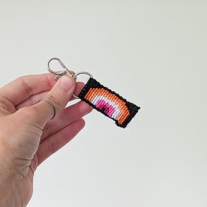 May include: A handmade keychain featuring a woven rainbow design in shades of orange, pink, and white, set against a black background. The keychain is attached to a silver-colored clasp. The item is held against a white background.