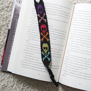 Puede incluir: Un marcapáginas de pulsera de la amistad tejido en negro con tres motivos apilados de calaveras y tibias cruzadas en morado, rojo y amarillo, con tibias correspondientes en rosa, naranja y azul claro, acabado con un lazo anudado y borla.