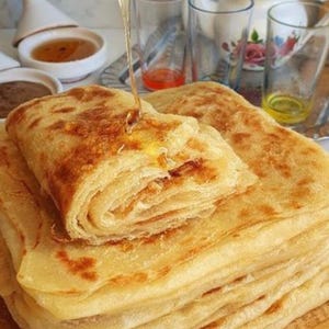 May include: A stack of golden-brown, flaky pastries, with a folded top piece, is drizzled with honey. Clear glasses, a teapot, and small bowls of honey and a dark spread are in the background, suggesting a breakfast or brunch setting.
