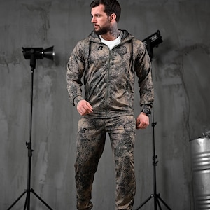 Könnte beinhalten: Ein zweiteiliger Sportanzug mit Tarnmuster in Braun-, Schwarz- und Beigetönen. Die Jacke hat eine Kapuze und einen Reißverschluss, die Hose ist gerade geschnitten. Das Outfit wird mit einem weißen T-Shirt und weißen Turnschuhen getragen.