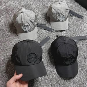 May include: Two silver-grey and two black baseball caps made of a crinkled nylon material. Each cap features a circular patch on the front panel with the text Stone Island and a compass logo. The caps include black tags attached to the back straps.