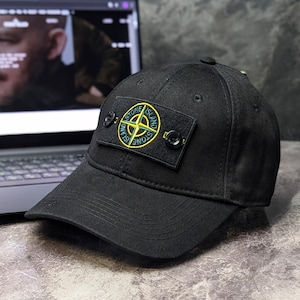 Könnte beinhalten: Eine schwarze Baseballkappe mit einem rechteckigen Aufnäher auf der Vorderseite. Der Aufnäher zeigt ein gelb-grünes Kompass-Logo mit dem Schriftzug Stone Island und ist mit zwei schwarzen Knöpfen neben dem gestickten Emblem befestigt.