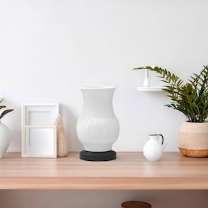May include: A white ribbed vase with a black base sits on a light wood table, alongside a white vase with a plant, a small white pitcher, and a potted plant. A white picture frame and a woven decorative item are also on the table.