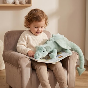 Puede incluir: Un juguete de peluche de dragón en color verde salvia con tela texturizada, alas pequeñas y una cola larga, descansando sobre un libro ilustrado abierto. El dragón presenta un hocico redondeado, orejas pequeñas y ojos oscuros.