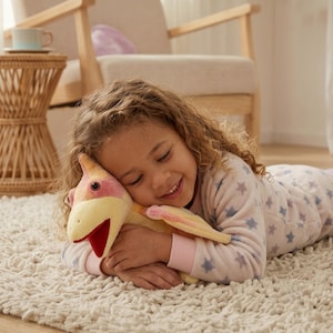 Peut inclure: Une peluche ptérodactyle avec un corps jaune doux, des accents d'ailes roses et une bouche ouverte rouge foncé. Le dinosaure en peluche présente une expression amicale avec des yeux sombres et est fabriqué dans un tissu duveteux.
