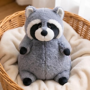Op de afbeelding: Een wasbeerknuffel met zachte grijze vacht, een wit gezichtsmasker en zwarte oogvlekken. Het knuffeldier heeft een rond, mollig lichaam, kleine zwarte pootjes en afgeronde oren met witte details aan de binnenkant, zittend in een rechtopstaande houding.