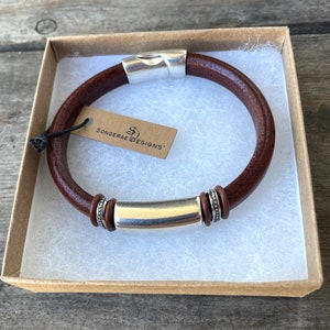 Pu&ograve; includere: Un braccialetto in pelle marrone con una barra in metallo argentato e dettagli decorativi. Il braccialetto &egrave; in una scatola con un'etichetta che recita "Sonserae Designs".