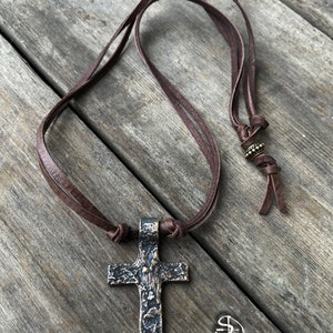 May include: A brown leather cord necklace with a large, textured, bronze cross pendant.