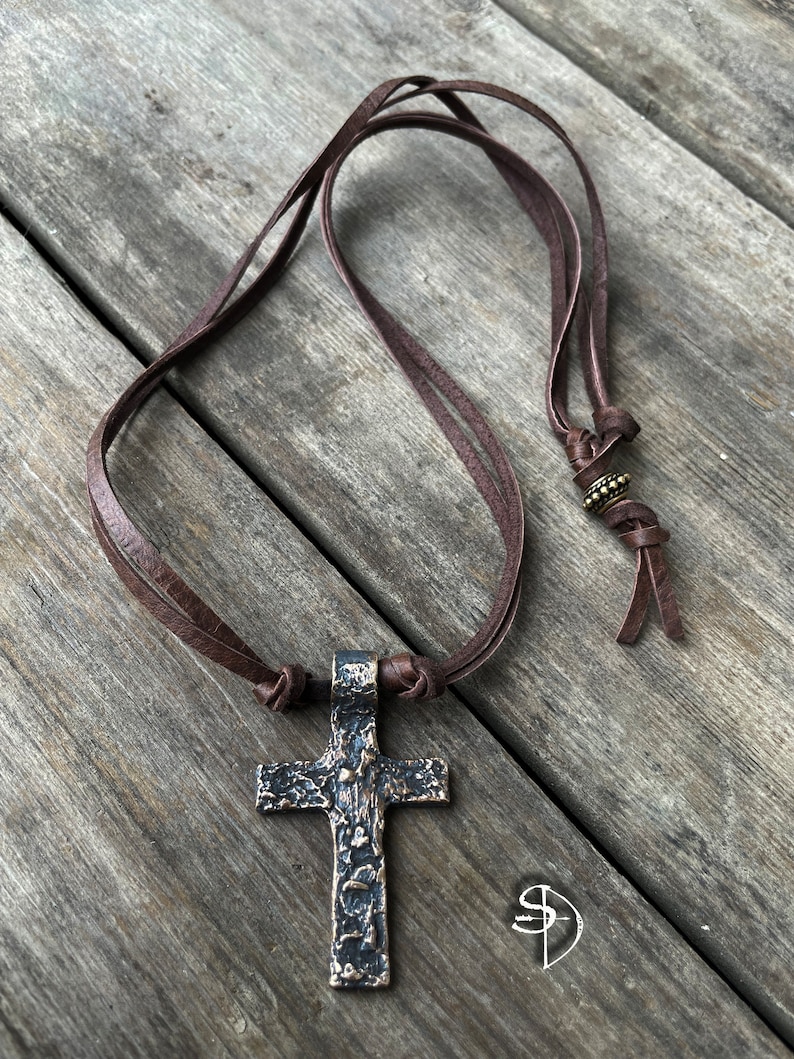 May include: A brown leather cord necklace with a large, textured, bronze cross pendant.