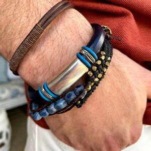 Pu&ograve; includere: Un set di braccialetti con una barra d'argento, perline blu, pelle marrone e dettagli in metallo dorato.