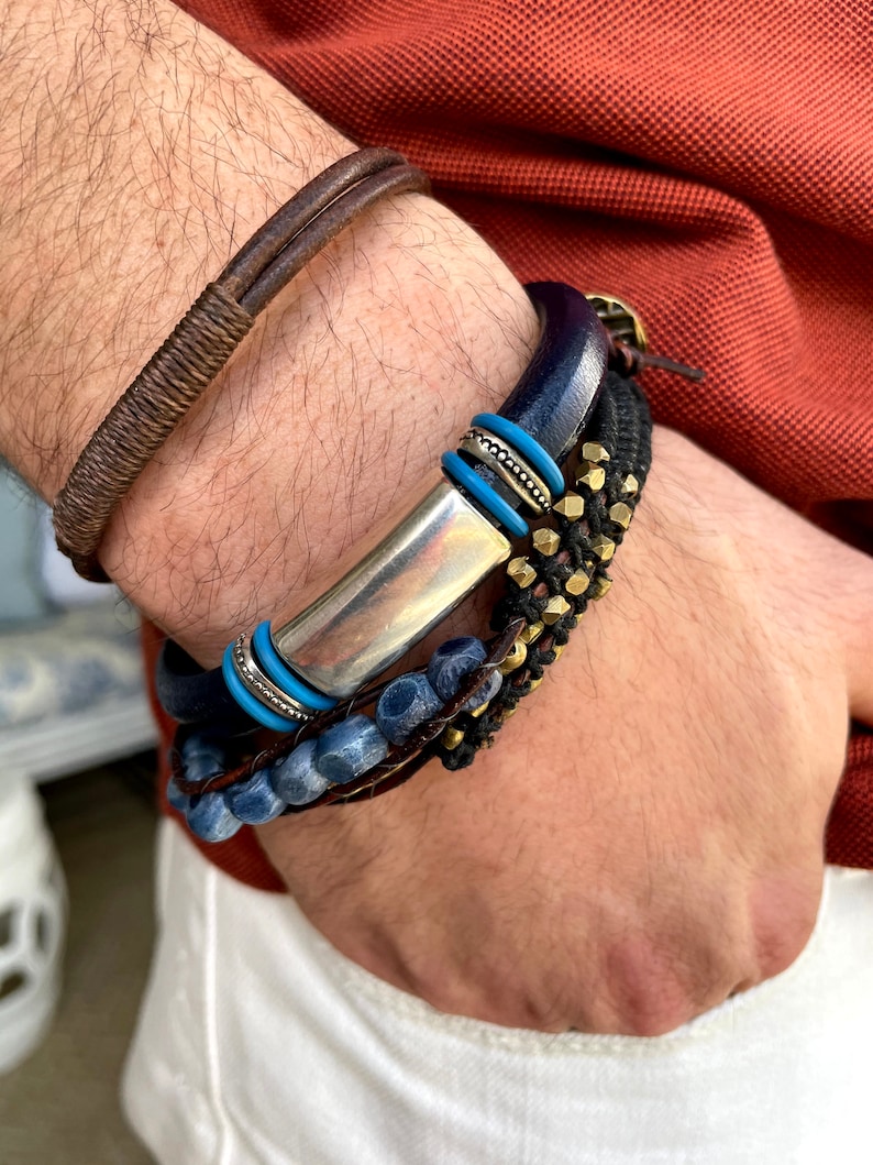 Pu&ograve; includere: Un set di braccialetti con una barra d'argento, perline blu, pelle marrone e dettagli in metallo dorato.