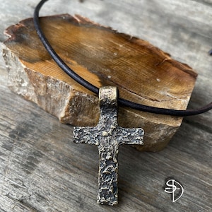 May include: A rustic, textured bronze cross pendant on a brown leather cord. The cross has a hammered finish and is a statement piece of jewelry.