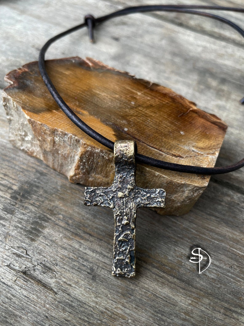 May include: A rustic, textured bronze cross pendant on a brown leather cord. The cross has a hammered finish and is a statement piece of jewelry.