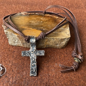 May include: A brown leather cord necklace with a hammered metal cross pendant. The cord has a decorative bead near the end.