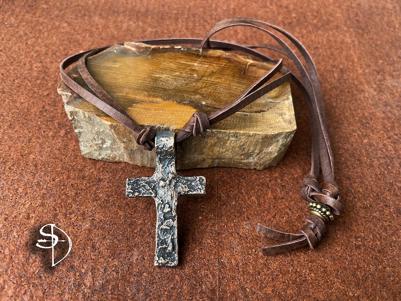 May include: A brown leather cord necklace with a hammered metal cross pendant. The cord has a decorative bead near the end.
