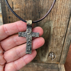 May include: A textured bronze cross pendant on a brown leather cord. The cross has a rustic, aged look.