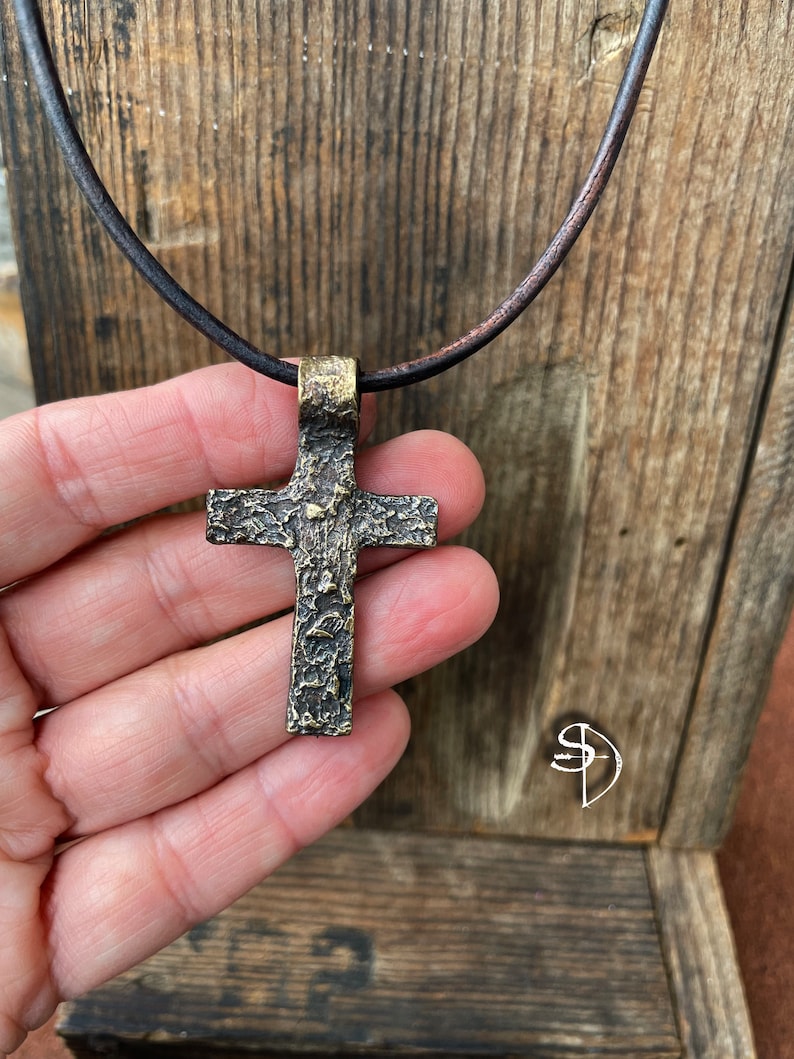 May include: A textured bronze cross pendant on a brown leather cord. The cross has a rustic, aged look.