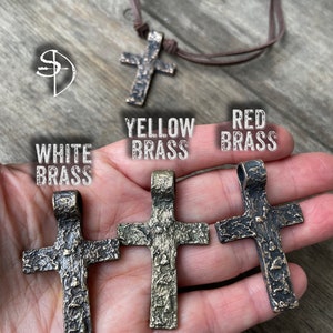 May include: Three metal cross pendants in different finishes: white brass, yellow brass, and red brass. Each cross has a textured surface and a loop at the top for hanging.