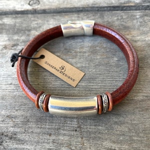 Pu&ograve; includere: Un braccialetto in pelle marrone con una barra in metallo argentato e anelli decorativi argentati. Il braccialetto ha una chiusura in metallo argentato e un'etichetta che recita "Sonserae Designs".