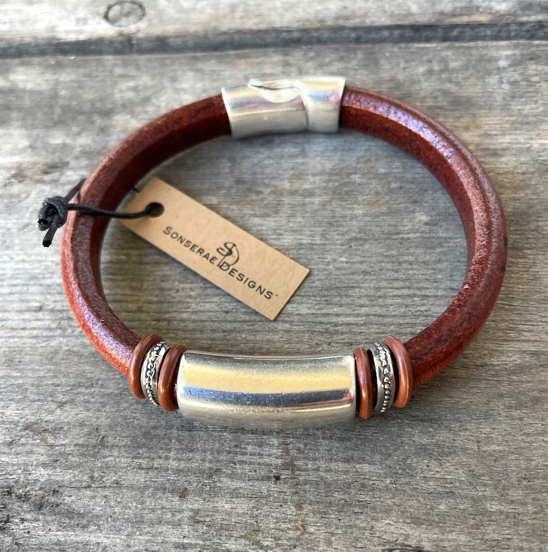 Pu&ograve; includere: Un braccialetto in pelle marrone con una barra in metallo argentato e anelli decorativi argentati. Il braccialetto ha una chiusura in metallo argentato e un'etichetta che recita "Sonserae Designs".