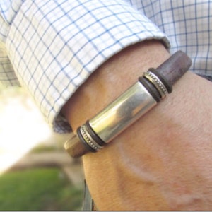 Pu&ograve; includere: Un braccialetto in pelle marrone con un ciondolo rettangolare argentato e anelli decorativi in argento.