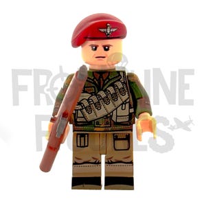 Op de afbeelding: Een Lego-figuur van een soldaat met een rode baret met een zilveren embleem, een camouflagejas en een beige broek. De figuur houdt een beige geweer vast. De soldaat heeft een serieuze uitdrukking.
