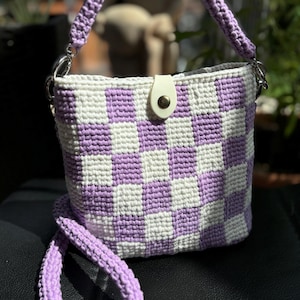 May include: A handmade crochet bag in a checkerboard pattern of white and lavender. The bag has a matching lavender strap and handle, with silver-tone metal hardware. A white leather tab with a gold snap closure secures the top.