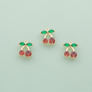 May include: Three gold-coloured cherry charms with red and green gemstone accents. Each charm features two red cherry-shaped stones and green leaf-shaped stones. The charms are set against a light green background.