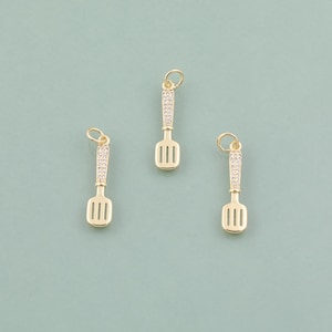 May include: Three gold-coloured spatula charms with a rhinestone-studded handle and a slotted head. Each charm has a small loop at the top for attaching to a necklace or bracelet. The charms are displayed on a light teal background.