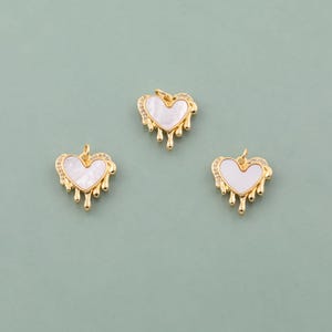Puede incluir: Tres colgantes dorados en forma de corazón con centros blancos y un diseño de goteo. Los colgantes tienen un halo de pequeñas piedras transparentes alrededor de la forma del corazón. El fondo es verde apagado.