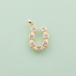 May include: A gold-colored horseshoe pendant with square, clear gemstones. The pendant has a loop at the top for a chain. The gemstones are set in a square shape and are evenly spaced along the horseshoe curve.