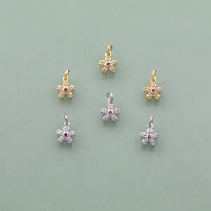 May include: Six flower-shaped charms. Some are gold-toned, others silver-toned. Each charm has a small, red center stone and is surrounded by clear, sparkling stones. The charms are designed for jewelry making or crafting.