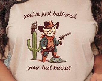 May include: Beige t-shirt featuring a cartoon cowboy cat with a gun, a cactus, and a bird. The text reads "you've just buttered your last biscuit."