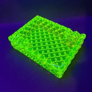 May include: A neon green ashtray with a diamond pattern. The ashtray is rectangular with rounded corners and has a raised edge. The ashtray is made of glass and has a glossy finish. The ashtray is on a dark blue surface.
