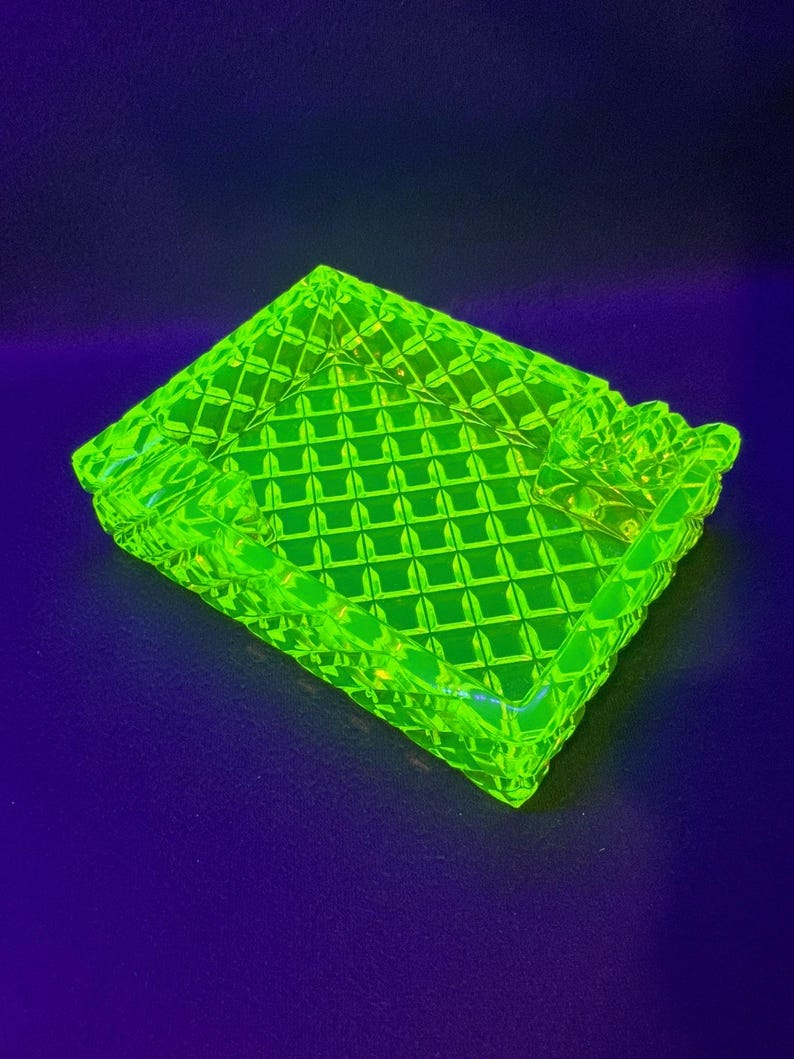 May include: A neon green ashtray with a diamond pattern. The ashtray is rectangular with rounded corners and has a raised edge. The ashtray is made of glass and has a glossy finish. The ashtray is on a dark blue surface.