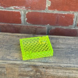 May include: A neon green, square-shaped trinket dish with a diamond pattern. The dish is made of a translucent material and sits on a wooden surface. The background features a red brick wall.