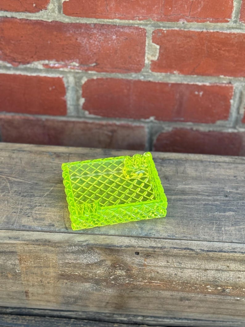May include: A neon green, square-shaped trinket dish with a diamond pattern. The dish is made of a translucent material and sits on a wooden surface. The background features a red brick wall.