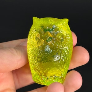 May include: A small, translucent, neon yellow glass owl figurine. The object features a textured surface with molded details representing feathers, two circular eyes, and pointed ears. The glass has a bright, vibrant, semi-transparent finish.