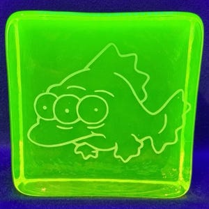 May include: A square, neon green glass coaster with a cartoon fish design. The fish has three eyes and a fin. The coaster is a vibrant, eye-catching accessory.