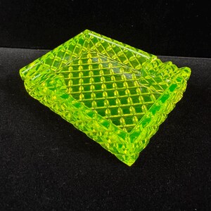 May include: A square, neon green glass ashtray with a diamond pattern. The ashtray has a raised, textured border and is set against a dark background. The glass has a bright, luminous quality.