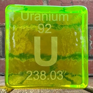 Op de afbeelding: Een vierkant, doorschijnend, neon groen en geel glazen blok met het woord "Uranium", het nummer "92", de letter "U" en het nummer "238.03" in het oppervlak geëtst. Het blok heeft een getextureerd, gelaagd uiterlijk.