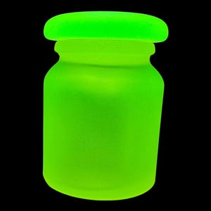Könnte beinhalten: Ein leuchtend neongrünes Glas mit passendem Deckel. Das Glas hat einen abgerundeten Körper und eine leicht strukturierte Oberfläche. Der Deckel sitzt fest obenauf und erzeugt ein zusammenhängendes, monochromes Design. Der Hintergrund ist tiefschwarz.