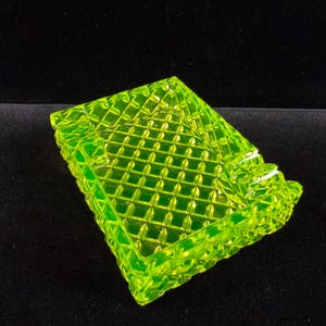 May include: A square, neon green glass ashtray with a diamond pattern. The edges of the ashtray have a decorative, scalloped design. The ashtray is made of a translucent glass material.