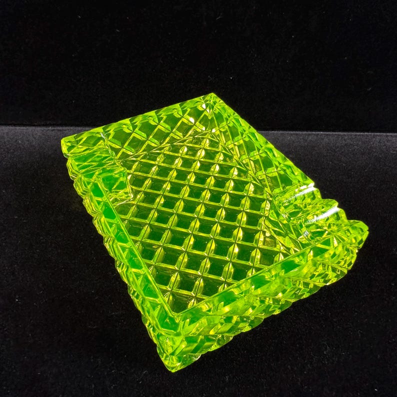 May include: A square, neon green glass ashtray with a diamond pattern. The edges of the ashtray have a decorative, scalloped design. The ashtray is made of a translucent glass material.