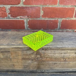 May include: A neon green, rectangular ashtray with a textured, grid-like pattern. The ashtray is made of a translucent material and sits on a weathered wooden surface, with a red brick wall in the background.