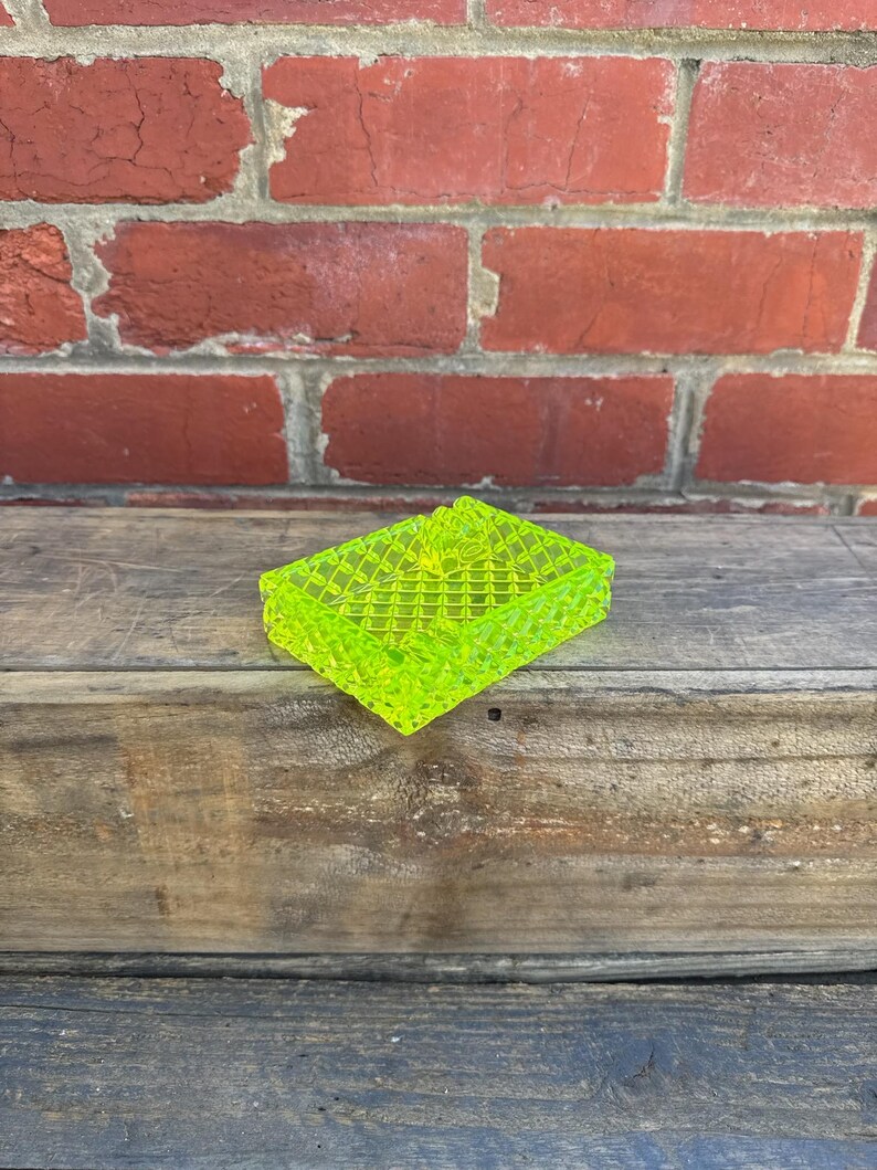 May include: A neon green, rectangular ashtray with a textured, grid-like pattern. The ashtray is made of a translucent material and sits on a weathered wooden surface, with a red brick wall in the background.