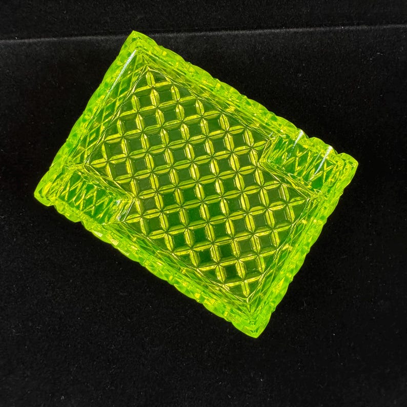 May include: A square, neon green glass ashtray with a diamond pattern. The ashtray has a textured, geometric design and a slightly scalloped edge. The ashtray is made of a translucent, bright green glass.